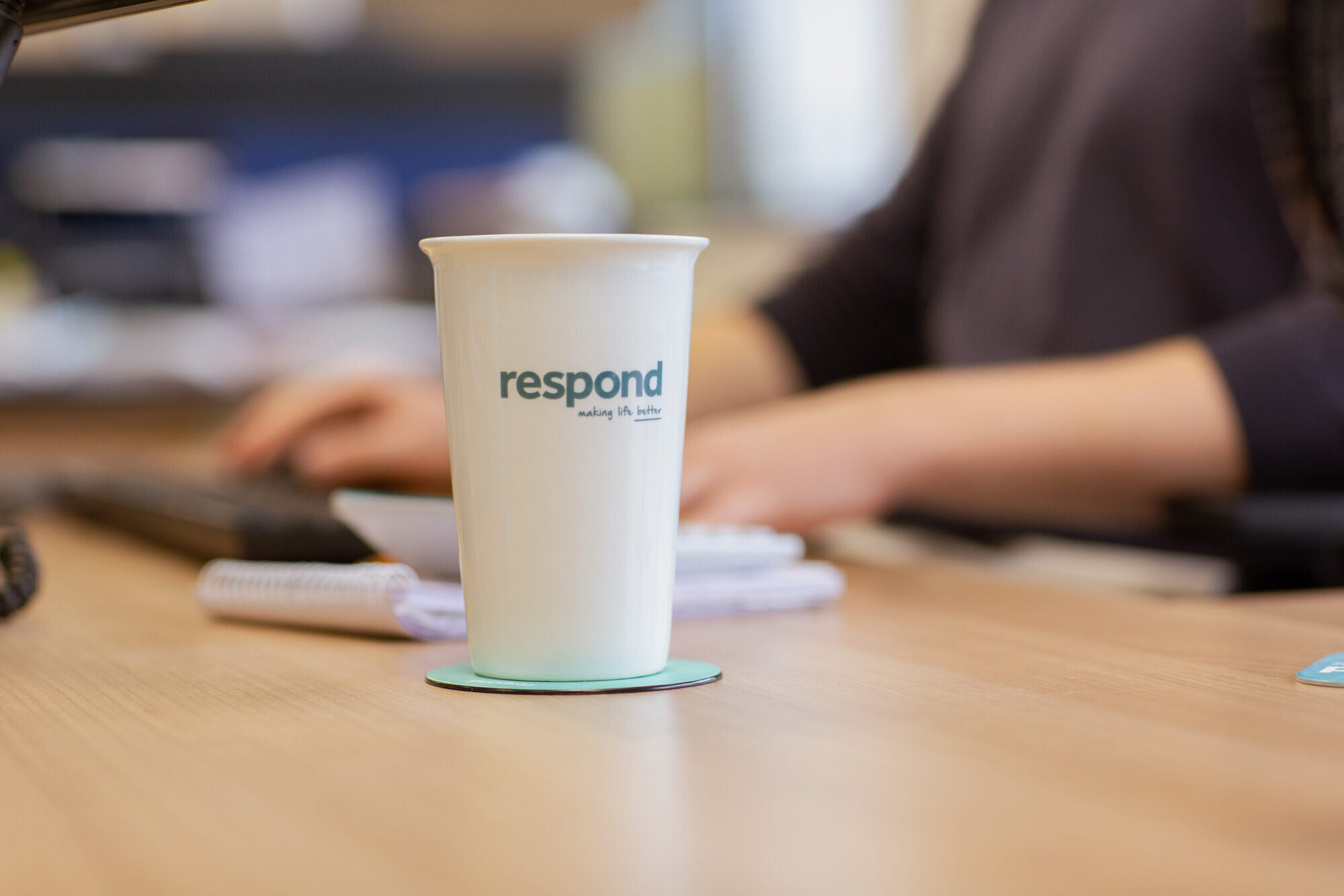 A Respond branded coffee cup sits on a desk with a blurred background of a Respond team member typing on a keyboard.