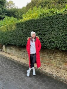 Amy, looking stylish wearing a red coat and white hat poses in front of a vibrant green hedge.